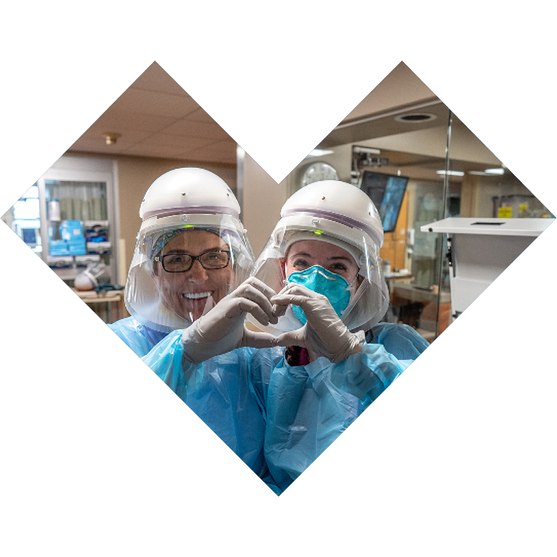 Two medical staff wearing protective clothing, gloves, and powered air-purifying respirators. They are holding their hands together to make a heart. 